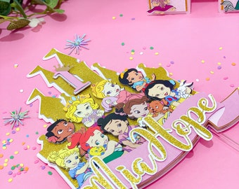 Può includere: Un topper per torta a forma di castello rosa e oro con un numero uno dorato e il nome Mia Hope. Il castello è decorato con vari personaggi delle principesse Disney, tra cui Cenerentola, Belle, Ariel e Biancaneve.