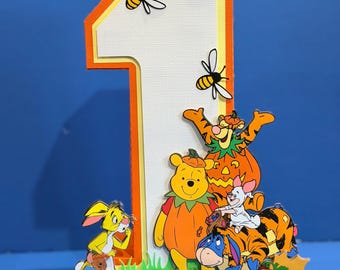 Può includere: Una grande decorazione tridimensionale a forma di numero uno con un centro bianco testurizzato e bordi arancioni. È decorata con due api e personaggi di Winnie the Pooh in costumi da zucca, con il nome MAXINE stampato in lettere arancioni.