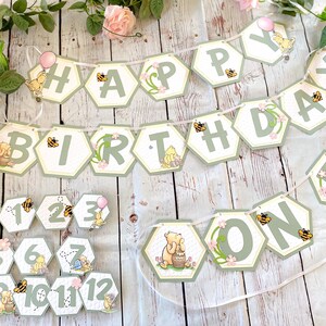 May include: A Winnie the Pooh themed birthday banner with green and yellow hexagonal pieces that spell out "Happy Birthday One". The banner is decorated with bees, flowers, and Winnie the Pooh characters. There are also 12 smaller hexagonal pieces with numbers 1 through 12 and the word "one" on them.