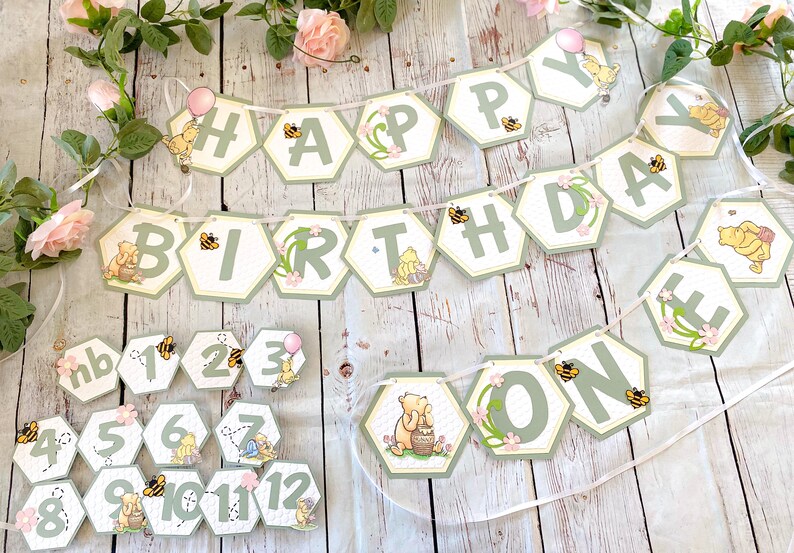 May include: A Winnie the Pooh themed birthday banner with green and yellow hexagonal pieces that spell out "Happy Birthday One". The banner is decorated with bees, flowers, and Winnie the Pooh characters. There are also 12 smaller hexagonal pieces with numbers 1 through 12 and the word "one" on them.