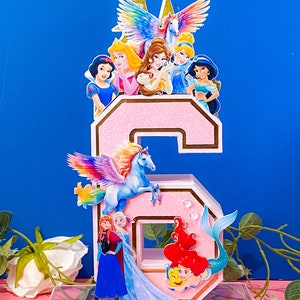 May include: A pink glitter number six cake topper with a variety of Disney princesses and characters, including Ariel, Elsa, Anna, Snow White, Cinderella, and a unicorn.