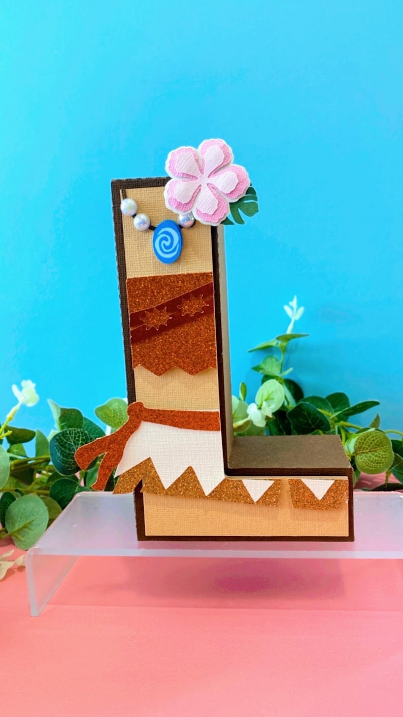 K&ouml;nnte beinhalten: Ein brauner Buchstabe L, verziert mit einer rosa Blume, einem blauen Wirbel und einer wei&szlig;en Perlenkette. Der Buchstabe steht auf einem durchsichtigen Acryl-St&auml;nder.