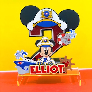 May include: A red number two cake topper with a Mickey Mouse character dressed as a captain. The topper includes a ship, a life preserver, and an anchor. The name "ELLIOT" is written in yellow letters on the ship.