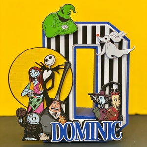 Puede incluir: Una letra de cart&oacute;n 'O' a rayas blancas y negras con un borde azul. La letra est&aacute; decorada con personajes de la pel&iacute;cula Pesadilla antes de Navidad, incluyendo a Jack Skellington, Sally, Oogie Boogie y Zero. La letra tambi&eacute;n est&aacute; decorada con el nombre "DOMINC" en letras azules.