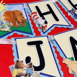 May include: A decorative banner featuring cream-colored pennants with scalloped blue edges, black letters, and illustrations of storybook characters including a brown bear, a cat in a striped hat, and a small bear sitting on a chair with a parent.