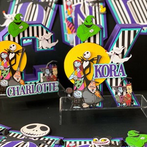 K&ouml;nnte beinhalten: Ein Kuchentopper zum Thema Nightmare Before Christmas mit den Buchstaben "KORA" und "CHARLOTTE" in Lila und Schwarz. Der Topper zeigt Figuren aus dem Film, darunter Jack Skellington, Sally und Oogie Boogie.