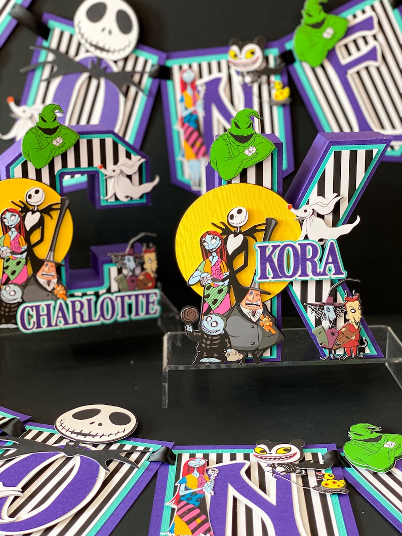 K&ouml;nnte beinhalten: Ein Kuchentopper zum Thema Nightmare Before Christmas mit den Buchstaben "KORA" und "CHARLOTTE" in Lila und Schwarz. Der Topper zeigt Figuren aus dem Film, darunter Jack Skellington, Sally und Oogie Boogie.