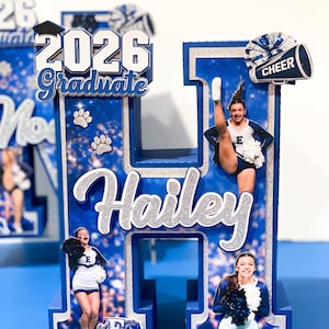 May include: A large 3D letter H decoration in blue and silver glitter, featuring the name Hailey in silver script, the text 2026 Graduate, and three photographs of a cheerleader in a blue and white uniform with pom-poms and a megaphone.