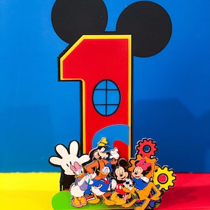 May include: A red number one cake topper with Mickey Mouse ears. The number one has a yellow outline and a blue window. Mickey Mouse, Minnie Mouse, Donald Duck, Goofy, Pluto, and a white glove are in front of the number one.