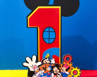 May include: A red number one cake topper with Mickey Mouse ears. The number one has a yellow outline and a blue window. Mickey Mouse, Minnie Mouse, Donald Duck, Goofy, Pluto, and a white glove are in front of the number one.