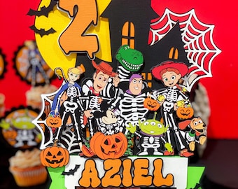 May include: A Halloween-themed cake topper featuring a black haunted house, a yellow moon with bats, and a group of skeleton characters from the Toy Story franchise. The characters include Woody, Buzz Lightyear, Jessie, Rex, and Alien. The topper also includes a pumpkin and the name "Aziel" in orange letters.
