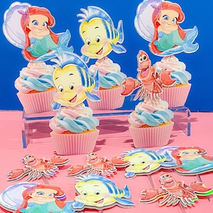 May include: A set of cupcake toppers featuring characters from The Little Mermaid. The toppers include Ariel, Flounder, and Sebastian. The toppers are printed on cardstock and have a glossy finish.