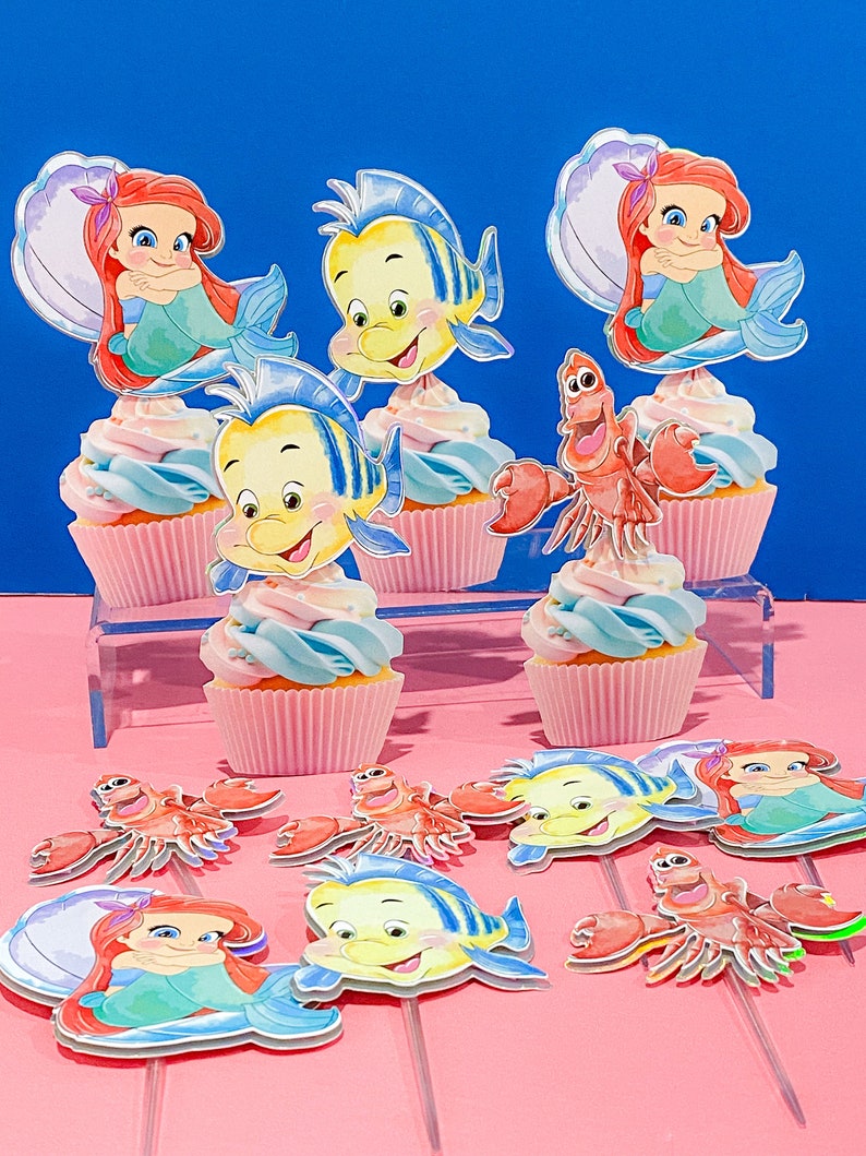 May include: A set of cupcake toppers featuring characters from The Little Mermaid. The toppers include Ariel, Flounder, and Sebastian. The toppers are printed on cardstock and have a glossy finish.