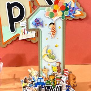 May include: A large, three-dimensional number one decoration featuring various storybook character cutouts, including a child holding balloons, a figure on a crescent moon, and a monkey reading a book. The base displays the name LEVI in bold letters.