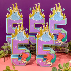 May include: Two purple and blue 3D number five cake toppers. Each number is decorated with a gold castle and a princess in a blue dress. The bottom of each number is decorated with a mermaid in a green tail.