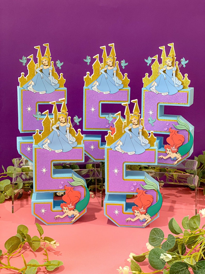 May include: Two purple and blue 3D number five cake toppers. Each number is decorated with a gold castle and a princess in a blue dress. The bottom of each number is decorated with a mermaid in a green tail.