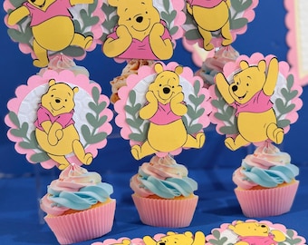Può includere: Una collezione di decorazioni per cupcake raffiguranti personaggi gialli di Winnie the Pooh con magliette rosa, su sfondi circolari smerlati rosa e bianchi testurizzati con accenti di foglie verdi, sopra cupcake con glassa a spirale pastello.