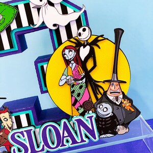 K&ouml;nnte beinhalten: Ein dreidimensionaler Kuchentopper in Form der Zahl drei, in den Farben Lila und T&uuml;rkis, auf einem schwarz-wei&szlig; gestreiften Hintergrund. Der Topper zeigt Figuren aus dem Film Nightmare Before Christmas, darunter Jack Skellington, Sally und Oogie Boogie. Der Topper enth&auml;lt auch den Namen "SLOAN" in lila und t&uuml;rkisfarbenen Buchstaben.