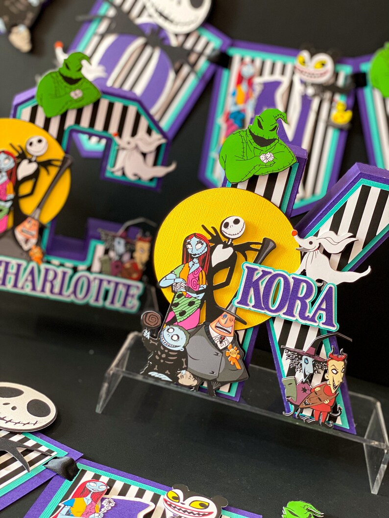 K&ouml;nnte beinhalten: Ein Kuchentopper zum Thema Nightmare Before Christmas mit den Namen Charlotte und Kora. Der Topper zeigt Figuren aus dem Film, darunter Jack Skellington, Sally und Oogie Boogie. Der Topper ist aus Papier und in den Farben Schwarz, Wei&szlig;, Lila, Gr&uuml;n und Gelb verziert.