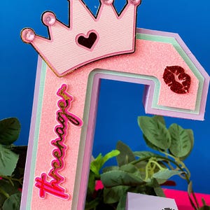 Puede incluir: Una letra 'T' de cart&oacute;n en capas de color rosa y verde con una corona rosa con purpurina dorada y un coraz&oacute;n rojo en la parte superior. La letra tiene la palabra 'teenager' escrita en purpurina dorada. La letra est&aacute; decorada con un labio rojo de purpurina y una bola de discoteca rosa y blanca.