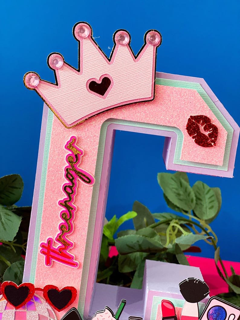 Puede incluir: Una letra 'T' de cart&oacute;n en capas de color rosa y verde con una corona rosa con purpurina dorada y un coraz&oacute;n rojo en la parte superior. La letra tiene la palabra 'teenager' escrita en purpurina dorada. La letra est&aacute; decorada con un labio rojo de purpurina y una bola de discoteca rosa y blanca.