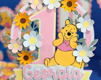 Può includere: Un topper per torta in carta a strati con un grande numero uno rosa all'interno di una cornice circolare decorata con fiori di carta bianchi, gialli e rosa. Un orso giallo con una maglietta rosa siede sulla base con il nome Georgia.