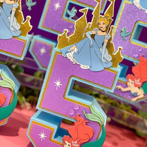 May include: Purple and blue glitter number five with a gold trim. The number has a cutout of Ariel the mermaid with red hair and a green tail. The number is decorated with gold stars and white glitter.