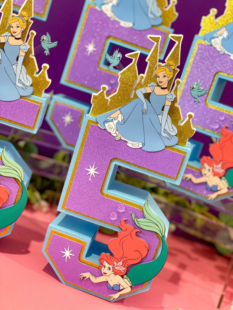 May include: Purple and blue glitter number five with a gold trim. The number has a cutout of Ariel the mermaid with red hair and a green tail. The number is decorated with gold stars and white glitter.
