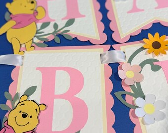 Può includere: Uno striscione decorativo composto da bandierine di carta con bordi smerlati e una trama a nido d'ape. Ogni bandierina presenta una grande lettera rosa, un personaggio di Winnie the Pooh giallo e decorazioni floreali in carta come girasoli e margherite.