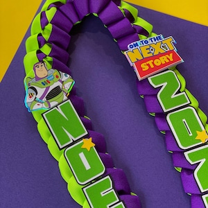 Op de afbeelding: Een groene en paarse lintlei met de woorden "NOEL" en "ZOE" gespeld in groene letters. Een cartoonafbeelding van Buzz Lightyear uit Toy Story is aan de lei bevestigd. Een kleine, kleurrijke sticker met de woorden "On to the Next Story" is ook aan de lei bevestigd.