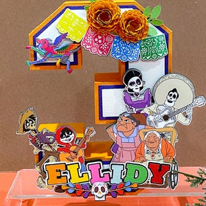 May include: A colorful 3D number "3" cake topper decorated with characters from the movie "Coco" and marigold flowers. The topper also includes the name "ELLIDY" in colorful letters and a sugar skull.