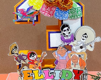May include: A colorful 3D number "3" cake topper decorated with characters from the movie "Coco" and marigold flowers. The topper also includes the name "ELLIDY" in colorful letters and a sugar skull.