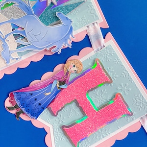 Może przedstawiać: Dekoracyjny baner z r&oacute;żową, falistą krawędzią. Zawiera ilustrację jasnoniebieskiego konia, r&oacute;żową literę "H" z brokatem i ilustrację postaci w niebieskiej sukience. Baner ma jasnoniebieskie tło ze wzorami płatk&oacute;w śniegu.