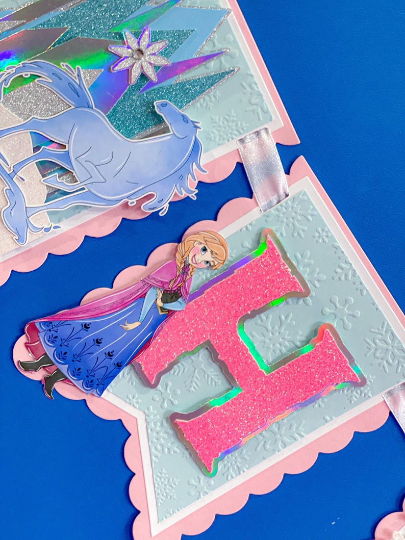 Może przedstawiać: Dekoracyjny baner z r&oacute;żową, falistą krawędzią. Zawiera ilustrację jasnoniebieskiego konia, r&oacute;żową literę "H" z brokatem i ilustrację postaci w niebieskiej sukience. Baner ma jasnoniebieskie tło ze wzorami płatk&oacute;w śniegu.