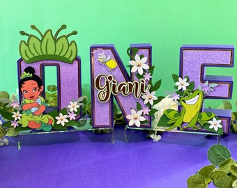 Könnte beinhalten: Lilafarbene Glitzerbuchstaben, die "ONE" buchstabieren, mit einer grünen Krone, einem Frosch und einer Prinzessin. Die Buchstaben sind mit Blumen und Blättern verziert. Der Name "Giani" ist in brauner Kursivschrift auf dem "O" geschrieben.