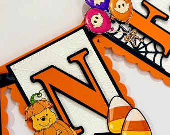 Può includere: Uno striscione a tema Halloween con la lettera N in arancione su sfondo bianco con trama a ragnatela. La lettera è decorata con un orso giallo in costume da zucca, due caramelle a forma di mais e un grappolo di palloncini a fantasma.