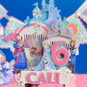 May include: A multi-layered cake topper featuring characters from the animated film "Frozen." The design includes a castle, a crown, and the number "6" in pink. The name "CALI" is displayed at the bottom, with a pink and white color scheme.