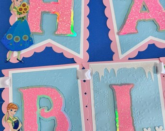 Può includere: Uno striscione decorativo con gagliardetti azzurri dalla trama a fiocchi di neve, bordi smerlati rosa e nastro bianco. Le lettere H, A, B e I sono in rosa glitterato con bordi olografici. Gli accenti includono motivi a girasole e figure animate.