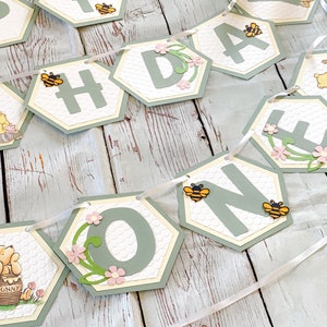May include: A green and white honeycomb-themed banner with the words "Happy 1st Birthday" and Winnie the Pooh characters. The banner is strung together with white ribbon.