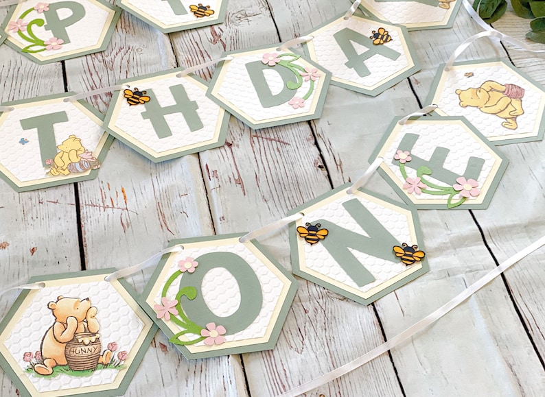 May include: A green and white honeycomb-themed banner with the words "Happy 1st Birthday" and Winnie the Pooh characters. The banner is strung together with white ribbon.