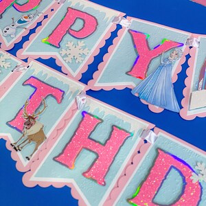 Może przedstawiać: Świąteczny baner z napisem "HAPPY BIRTHDAY" w r&oacute;żowych, brokatowych literach. Baner zawiera obrazy postaci i płatk&oacute;w śniegu na jasnoniebieskich proporczykach z r&oacute;żowymi, falistymi krawędziami. Srebrna korona wieńczy pierwszy proporczyk.