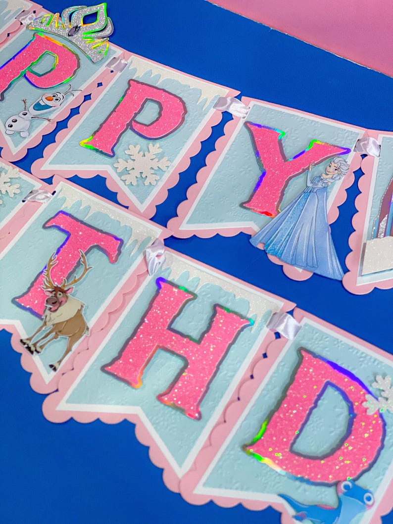 Może przedstawiać: Świąteczny baner z napisem "HAPPY BIRTHDAY" w r&oacute;żowych, brokatowych literach. Baner zawiera obrazy postaci i płatk&oacute;w śniegu na jasnoniebieskich proporczykach z r&oacute;żowymi, falistymi krawędziami. Srebrna korona wieńczy pierwszy proporczyk.