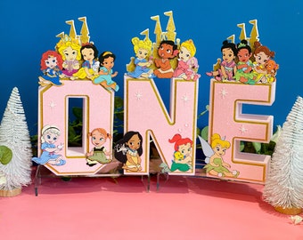 May include: A pink "ONE" birthday decoration with gold trim, featuring princess characters and castle toppers. The letters are decorated with glitter and star accents, perfect for a first birthday celebration.