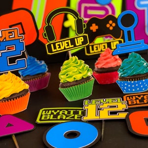 Pode incluir: Um conjunto de toppers para cupcakes com tema de videogame. Os toppers incluem um joystick, fones de ouvido, um controle de jogo e o texto "Level Up" e "Level 12".