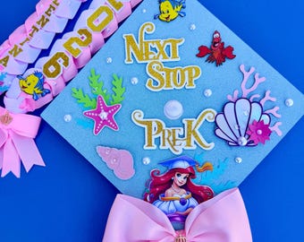 Può includere: Un topper per tocco di laurea azzurro decorato con scritte glitterate dorate che recitano Next Stop PreK, con sagome di una sirena, un pesce giallo, un granchio rosso, corallo rosa e conchiglie, arricchito da perle bianche e un grande fiocco rosa.