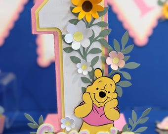Può includere: Una decorazione tridimensionale a forma di numero uno con bordo stratificato rosa e giallo, decorata con girasoli, margherite bianche e fogliame verde in carta. È applicato un orso giallo con maglietta rosa e il nome Georgia in rosa.