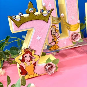 May include: Two pink and yellow foam letters, "Z" and "U", decorated with a gold crown, a gold ribbon, a gold-colored character, and a pink rose. The "Z" has a picture of Belle from Beauty and the Beast.
