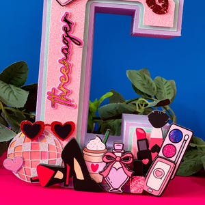 Puede incluir: Una letra C rosa con una corona en la parte superior y la palabra "Forever" escrita en el lateral. La letra est&aacute; decorada con varios art&iacute;culos femeninos, como una bola de discoteca, tacones altos, una taza de caf&eacute;, perfume, l&aacute;piz labial y un tel&eacute;fono.