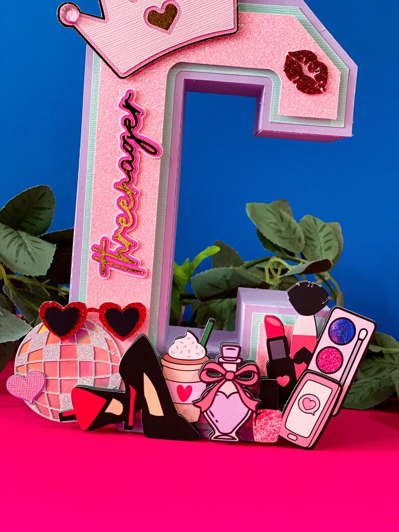 Puede incluir: Una letra C rosa con una corona en la parte superior y la palabra "Forever" escrita en el lateral. La letra est&aacute; decorada con varios art&iacute;culos femeninos, como una bola de discoteca, tacones altos, una taza de caf&eacute;, perfume, l&aacute;piz labial y un tel&eacute;fono.