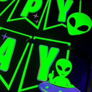 May include: A black banner with neon green text that reads "HAPPY BIRTHDAY". The banner features alien and UFO designs. The letters and alien figures glow brightly against the dark background, creating a vibrant, eye-catching effect.